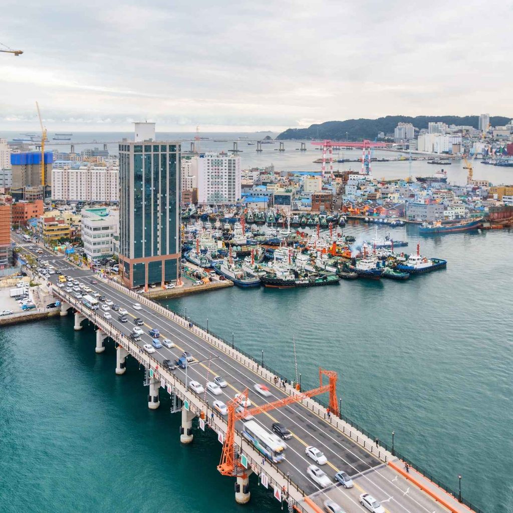 High-angle view of cars driving across Yeongdo Bridge with a busy harbor, ships, and cityscape in the background in Busan, South Korea.