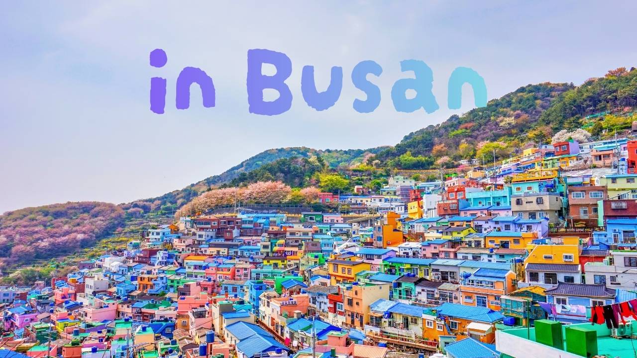 Colorful houses cascading down the hillside of Gamcheon Culture Village in Busan, with the text 'in Busan' overlaid in the sky.