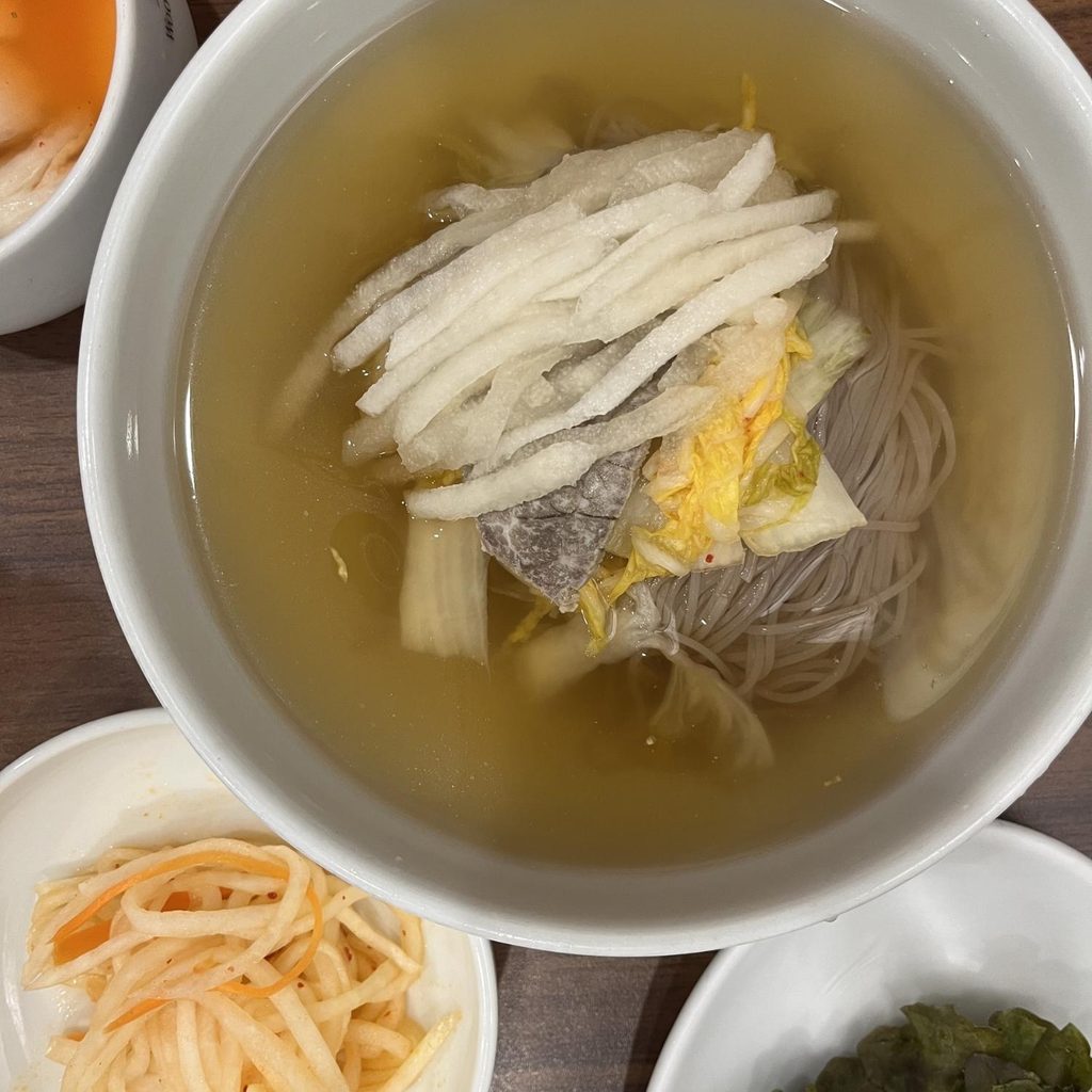 Pyongyang Naengmyeon bowl topped with sliced pears alongside traditional Korean marinated beef Bulgogi copper grill