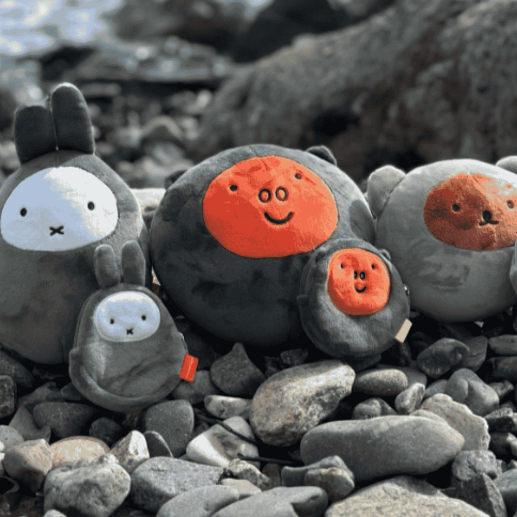 A collection of Geoje-exclusive gray Mongdol pebble-textured Miffy plush toys and round round pouches displayed naturally among real smooth pebbles on a coastal beach in South Korea.